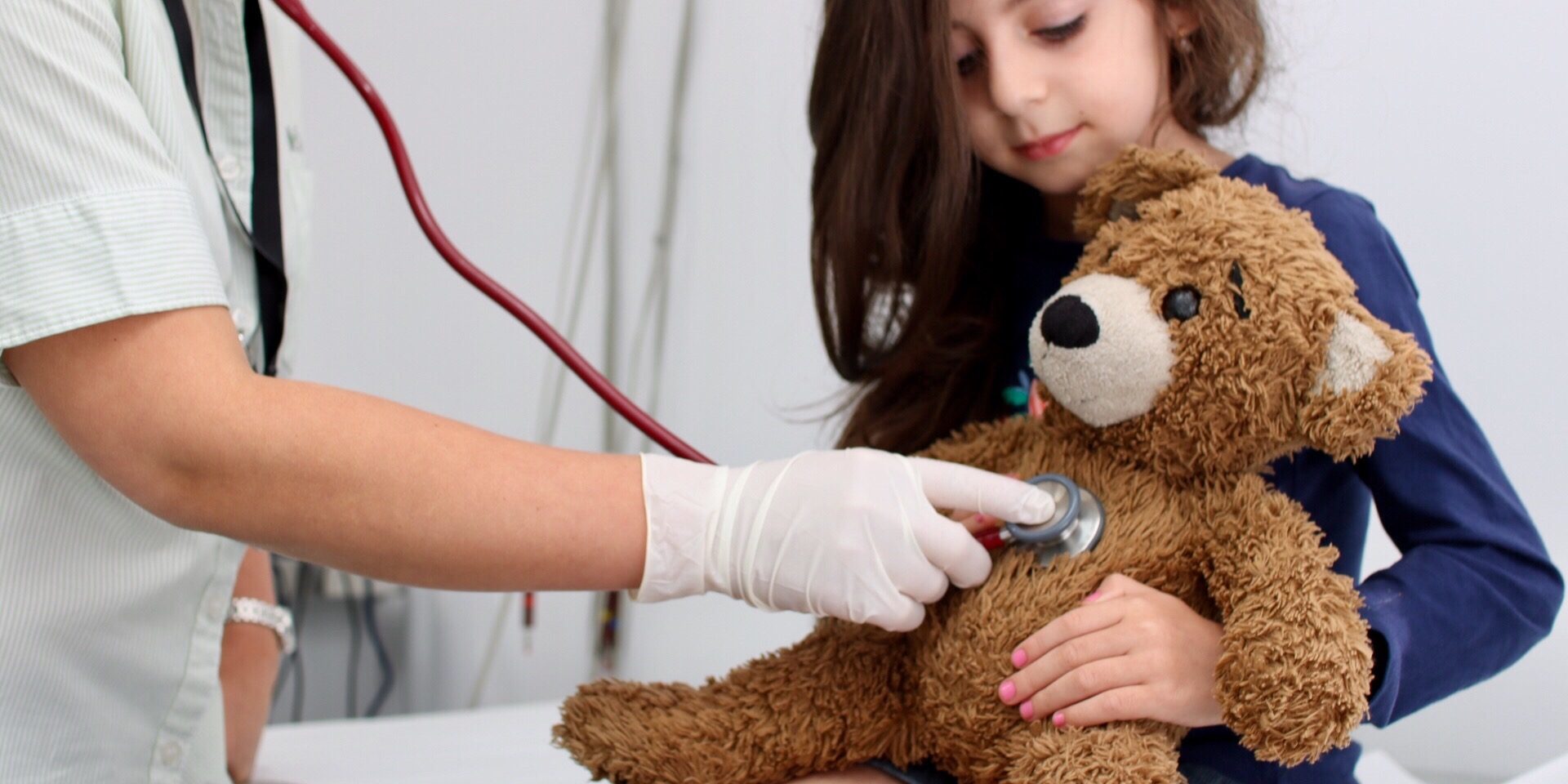 Medical professional using stethoscope on teddy bear in little girl's arms